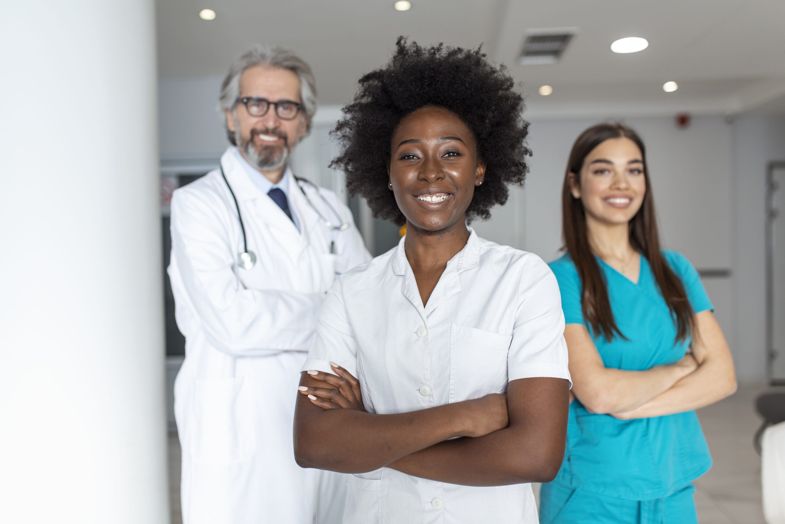 a multi ethnic group of three doctors and nurses standing in a hospital corridor, wearing scrubs and coats. the team of healthcare workers are staring at the camera and smiling